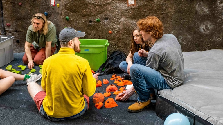 Members sitting on the ground near the climbing wall sorting through orange climbing holds.