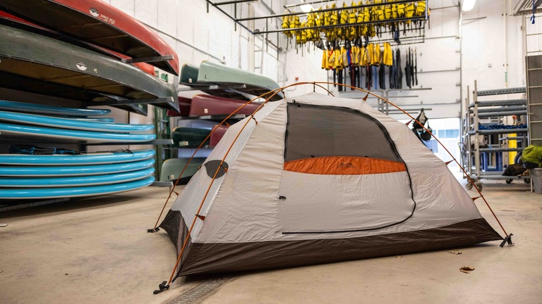 A tent is set up in the open area of the Outdoor Rental Center. There is equipment on all sides of the tent. The tent is grey with orange accents on the zippers and edges.