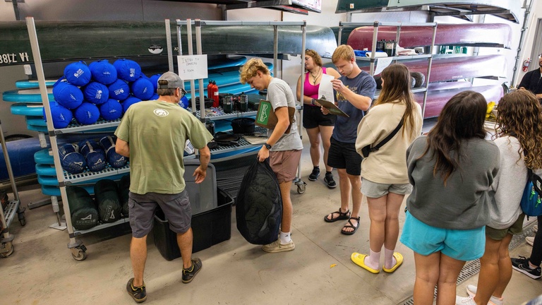 A group of students standing in the Outdoor Rental Center, they are all holding various pieces of equipment preparing to leave for an activity.