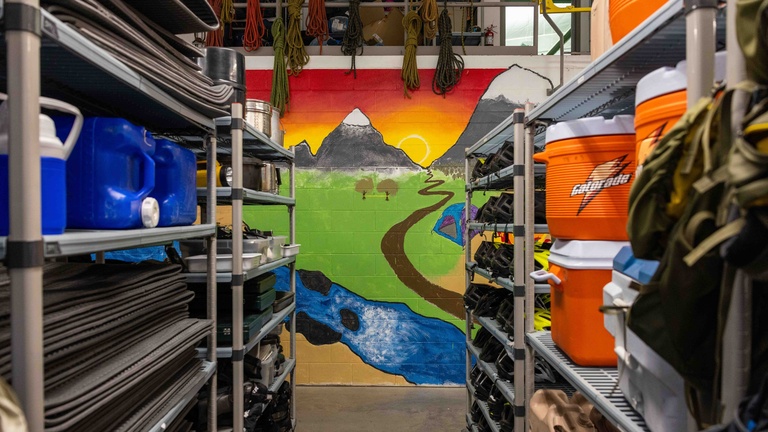 A photo looking at two shelving units full of equipment on either side of the frame. There is an aisle between the shelves and you can see a wall with a brightly colored mural of a mountain.