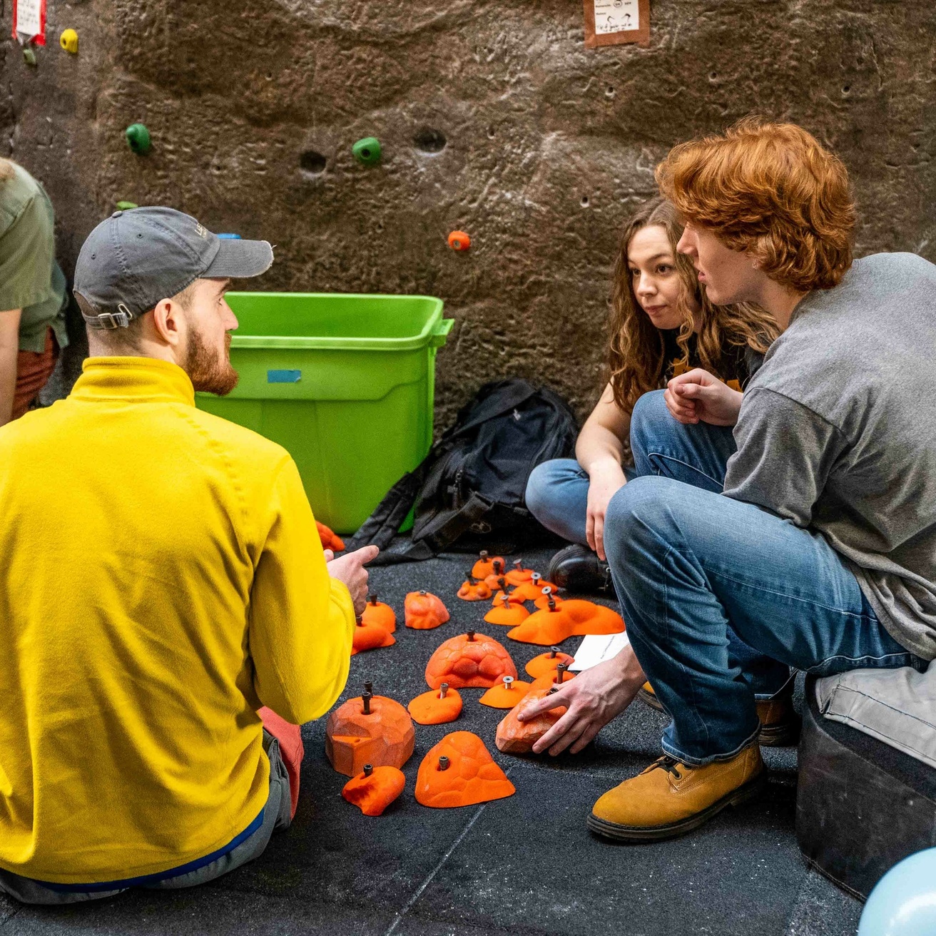 Members sitting on the ground near the climbing wall sorting through orange climbing holds.