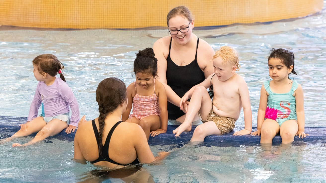 A group of four children sitting in the recreation pool. Two swim instructors are near them talking to the group.