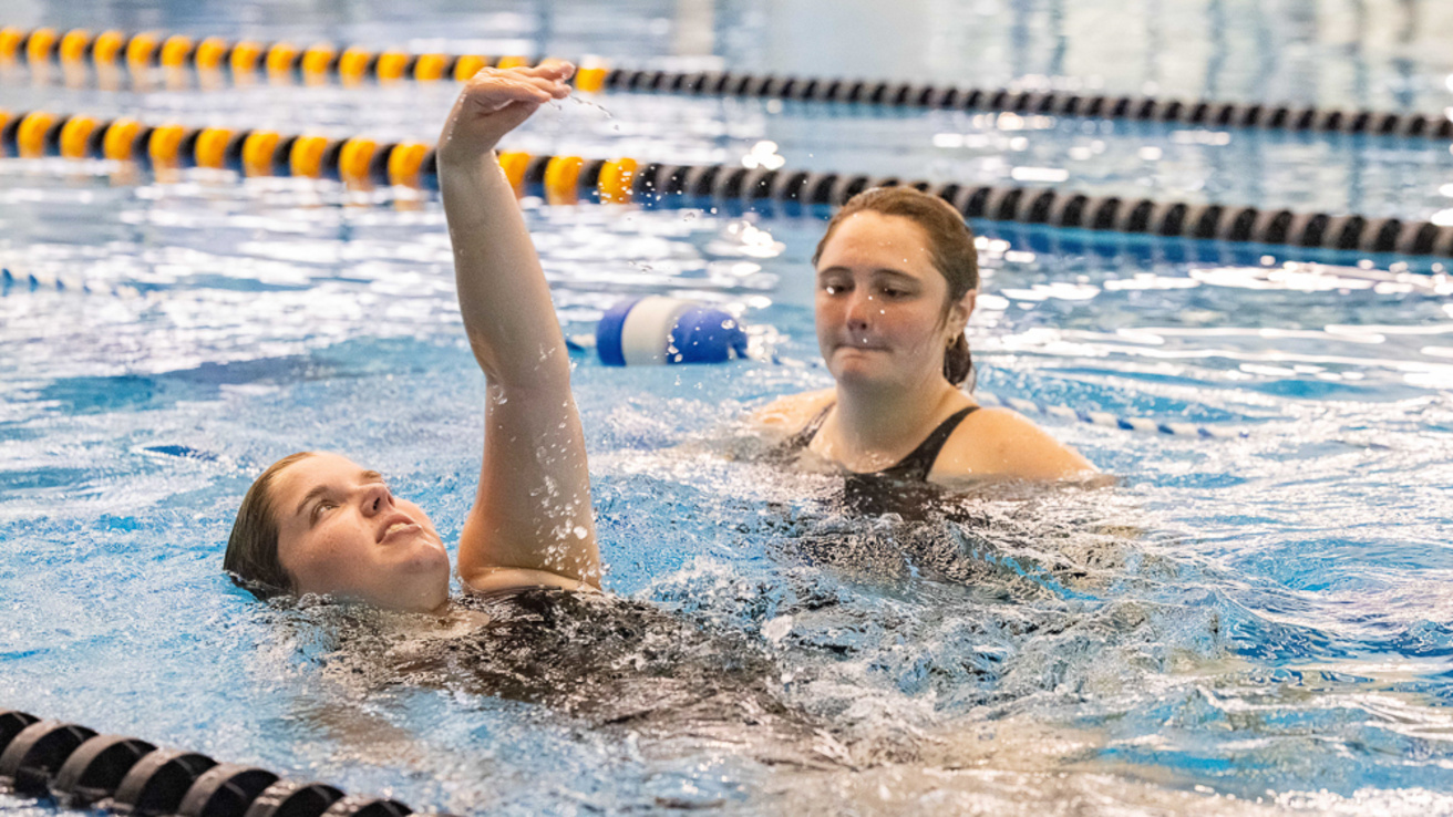 An adult swim lesson participant swimming on their back, hand in the air mid-stroke. An adult swim lesson instructor is next to them observing.
